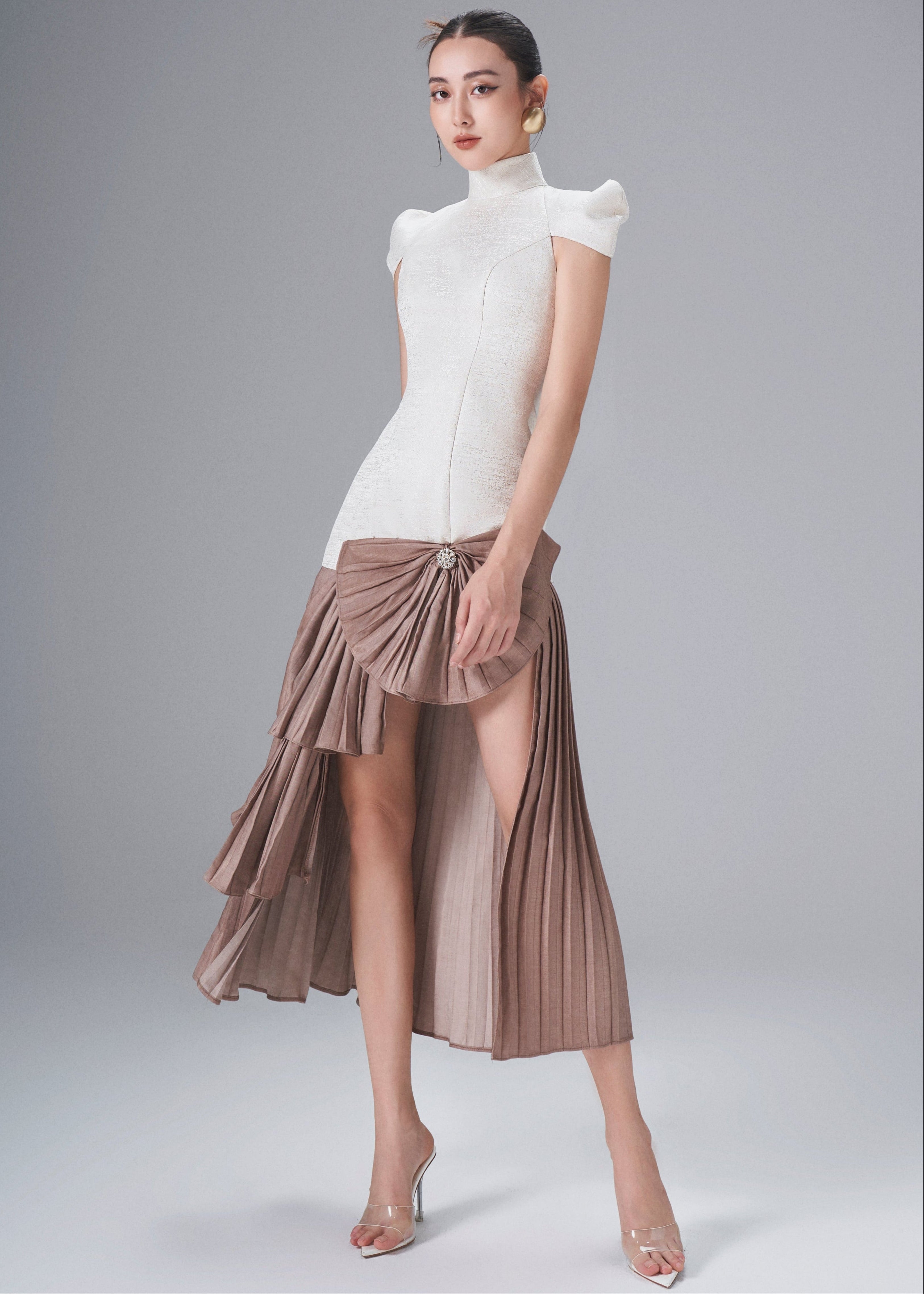 Woman wearing a white top and brown pleated skirt on a gray background